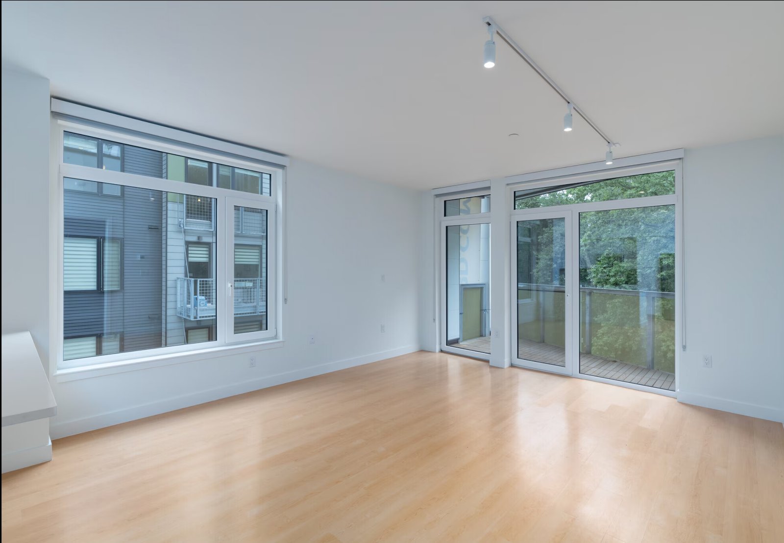 Empty living room, hardwood floor, large windows and balcony doors, no people